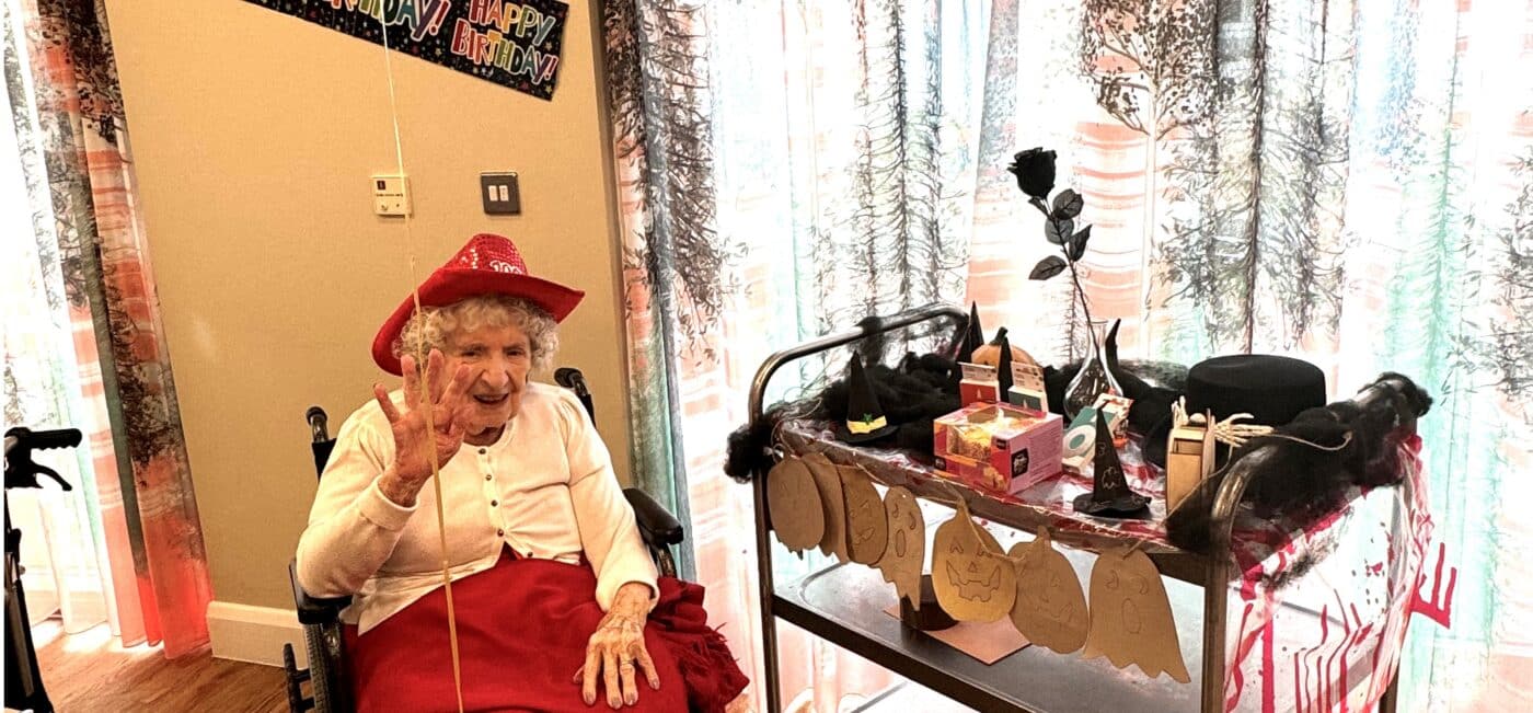 Older woman in red dress and hat waves cheerfully, seated by a table in a care home. A festive "Happy Birthday" banner hangs above. Decorated cart nearby.