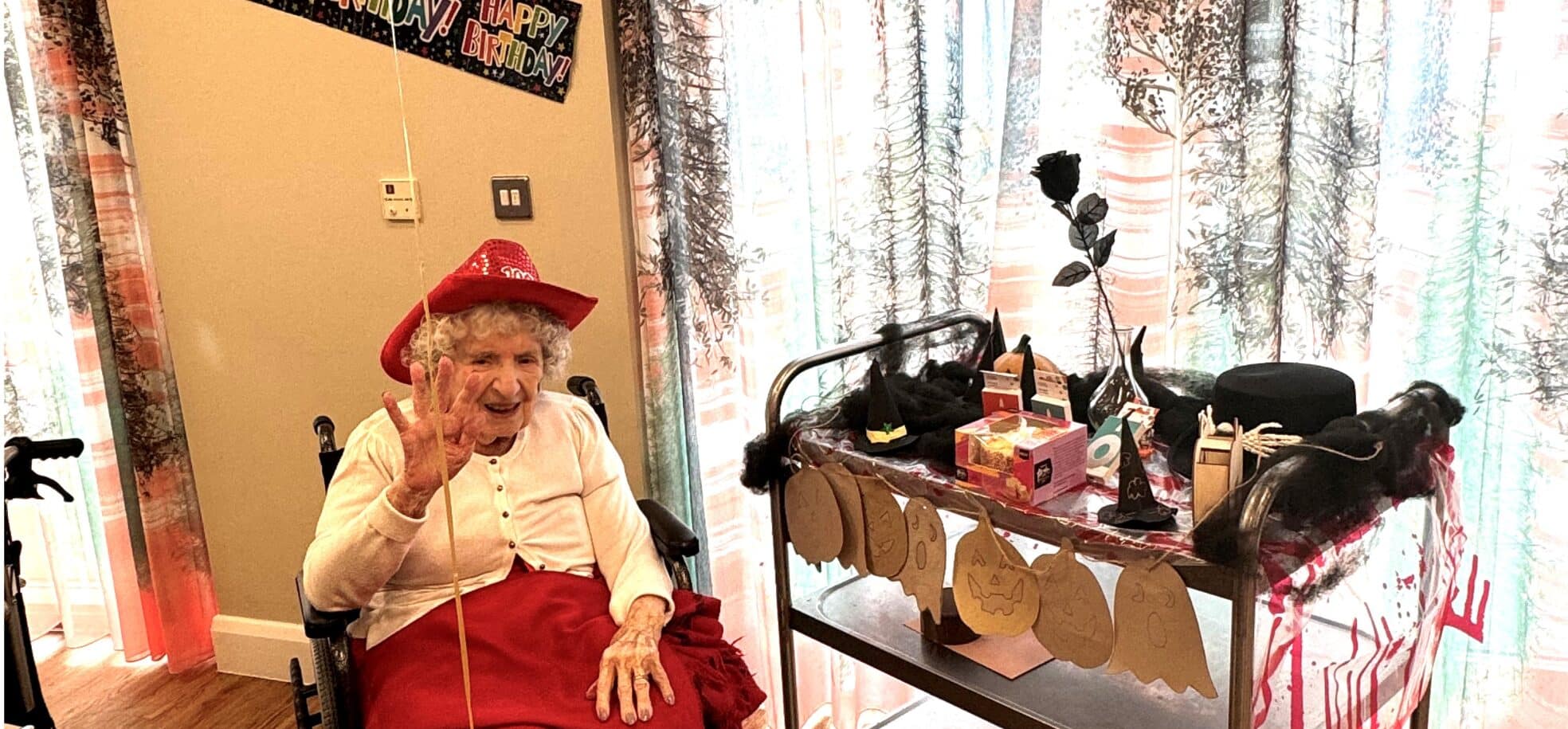 Older woman in red dress and hat waves cheerfully, seated by a table in a care home. A festive "Happy Birthday" banner hangs above. Decorated cart nearby.
