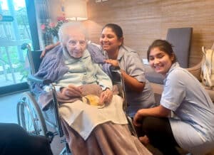 A care home resident smiling, eating cake and sitting in a wheelchair. Two staff members smiling next to her.