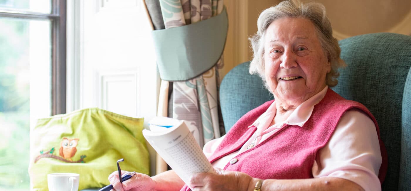 A care home resident, sitting in an armchair, smiles while doing a crossword.