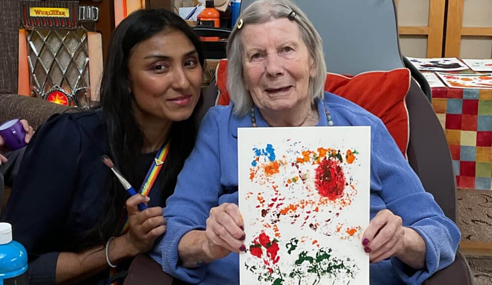 A care home resident holding up her creation, next to a staff member.