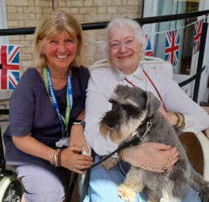 Two women, a volunteer and a resident, with a dog in the resident's lap.